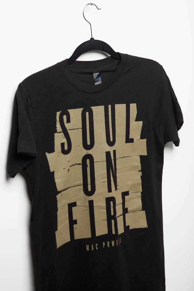 Close up of a black Mac Powell t-shirt with a gold design that reads "Soul of Fire".